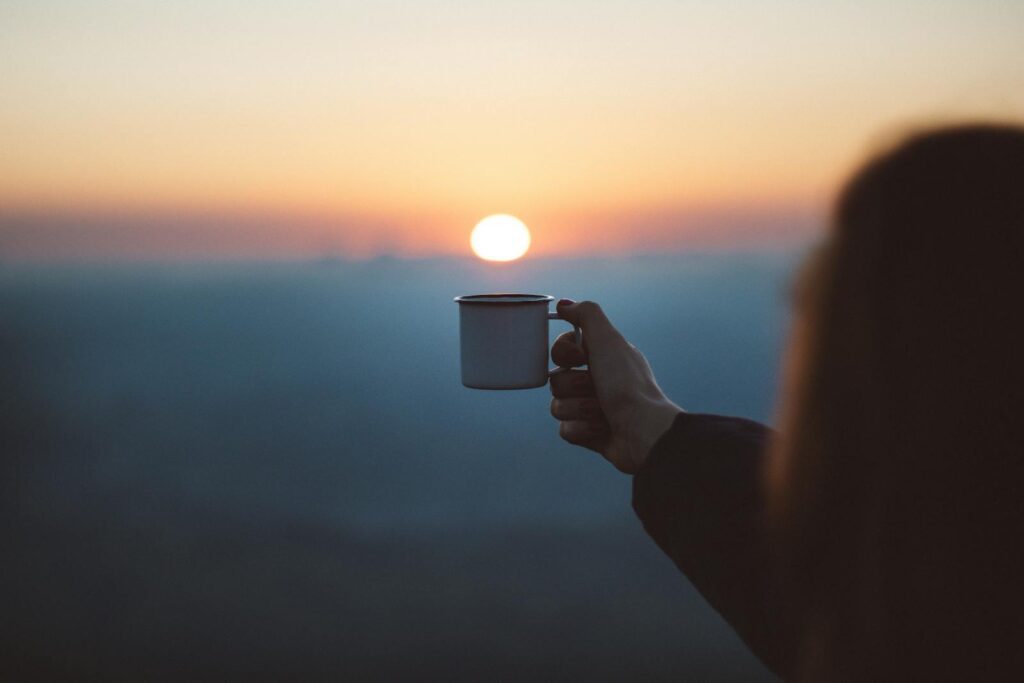Levantar Cedo: 4 Estratégias para Alcançar essa Meta Uma pessoa segurando uma caneca de café ao ar livre com o sol nascendo ao fundo. Levantar cedo.